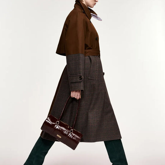 A model in side profile wearing a brown double-breasted coat over a dark brown plaid skirt, paired with dark green knee-high boots and carrying a dark brown handbag, set against a plain light gray studio background.