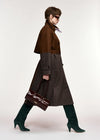 A model in side profile wearing a brown double-breasted coat over a dark brown plaid skirt, paired with dark green knee-high boots and carrying a dark brown handbag, set against a plain light gray studio background.