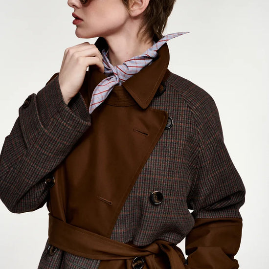 A model wearing a brown and gray houndstooth trench coat with a solid brown waist belt, a pink striped neck scarf, and dark sunglasses, photographed against a light gray studio background.