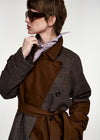 A model wearing a brown and gray houndstooth trench coat with a solid brown waist belt, a pink striped neck scarf, and dark sunglasses, photographed against a light gray studio background.