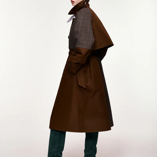 A model wearing a long brown coat with a structured dark gray shoulder cape, dark green suede knee-high boots, and black sunglasses, photographed against a plain white studio background.