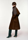 A model wearing a long brown coat with a structured dark gray shoulder cape, dark green suede knee-high boots, and black sunglasses, photographed against a plain white studio background.