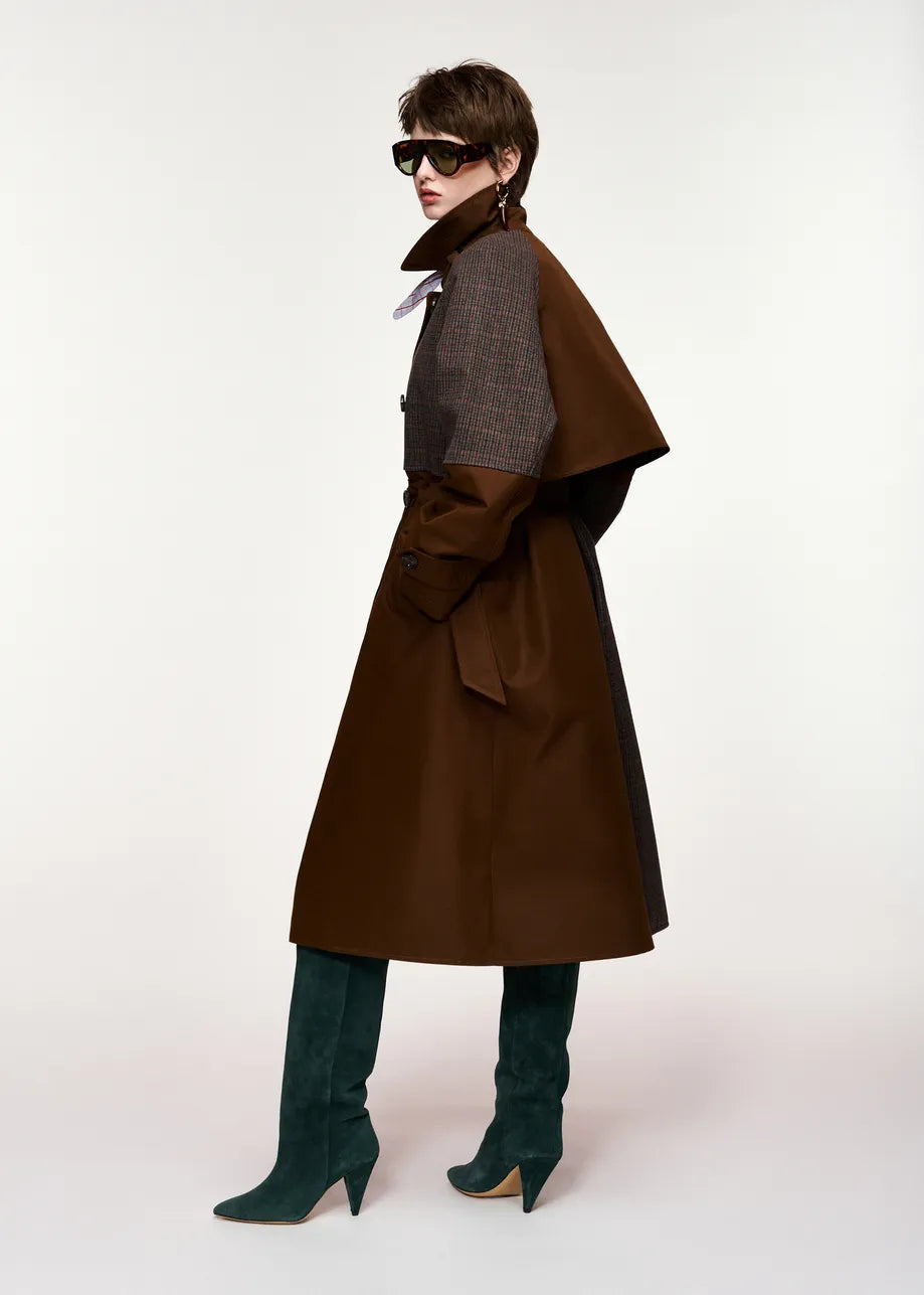 A model wearing a long brown coat with a structured dark gray shoulder cape, dark green suede knee-high boots, and black sunglasses, photographed against a plain white studio background.