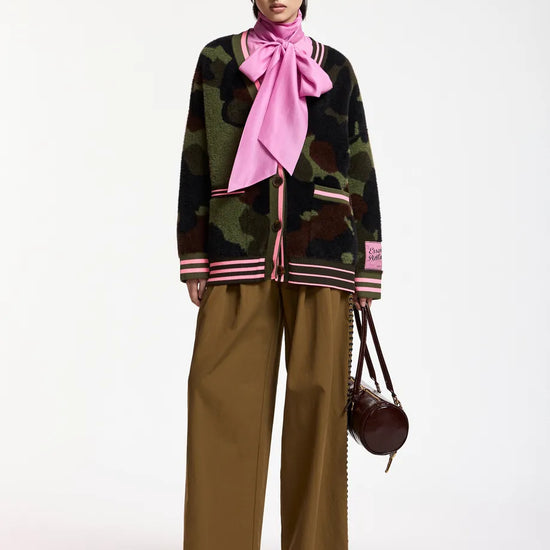 A model stands against a plain white studio background wearing a dark camouflage knit cardigan with pink striped trim, a large pink neck scarf, wide-leg brown pants, and black shoes, while holding a small dark brown handbag.