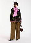 A model stands against a plain white studio background wearing a dark camouflage knit cardigan with pink striped trim, a large pink neck scarf, wide-leg brown pants, and black shoes, while holding a small dark brown handbag.