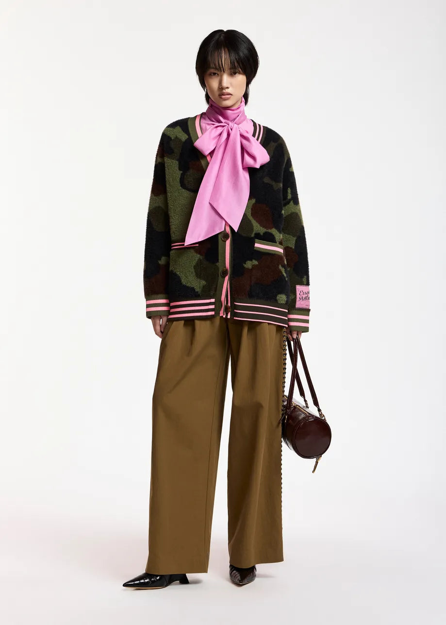 A model stands against a plain white studio background wearing a dark camouflage knit cardigan with pink striped trim, a large pink neck scarf, wide-leg brown pants, and black shoes, while holding a small dark brown handbag.