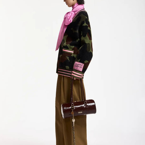 A model is shown from the side wearing a black camouflage jacket with pink and white striped ribbed cuffs and hem, paired with olive brown wide-leg trousers, a pink scarf, and carrying a dark brown cylindrical shoulder bag with a chain strap.