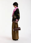 A model is shown from the side wearing a black camouflage jacket with pink and white striped ribbed cuffs and hem, paired with olive brown wide-leg trousers, a pink scarf, and carrying a dark brown cylindrical shoulder bag with a chain strap.