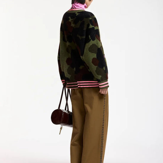 A model shown from the back wearing a green camouflage knit sweater with pink and white striped ribbed cuffs and hem, paired with wide-leg brown trousers and carrying a small dark red handbag, against a plain white studio background.