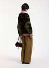 A model shown from the back wearing a green camouflage knit sweater with pink and white striped ribbed cuffs and hem, paired with wide-leg brown trousers and carrying a small dark red handbag, against a plain white studio background.