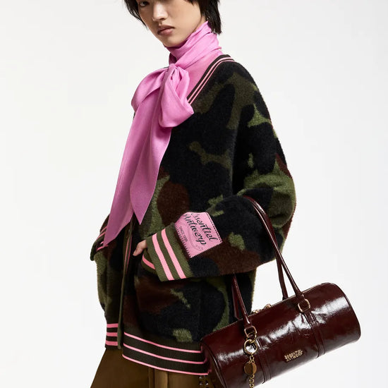A model wearing a dark camouflage knit cardigan with pink and white striped ribbed trim, a bright pink satin bow tie-neck scarf, and brown trousers, while carrying a dark brown cylindrical leather handbag.
