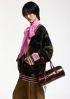 A model wearing a dark camouflage knit cardigan with pink and white striped ribbed trim, a bright pink satin bow tie-neck scarf, and brown trousers, while carrying a dark brown cylindrical leather handbag.
