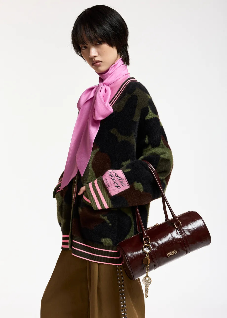 A model wearing a dark camouflage knit cardigan with pink and white striped ribbed trim, a bright pink satin bow tie-neck scarf, and brown trousers, while carrying a dark brown cylindrical leather handbag.