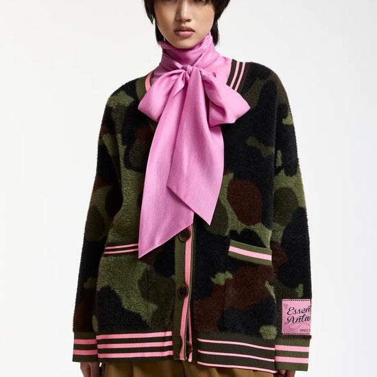 A model wearing a green and brown camouflage knit cardigan with pink striped trim, a large pink satin neck bow, and tan trousers, photographed against a plain white studio background.