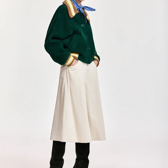 A model stands against a light gray studio background wearing a dark green corduroy jacket with gold and beige trim, a cream corduroy midi skirt, black knee-high boots, a blue neck scarf, and a beige baseball cap.