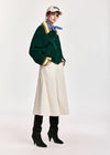 A model stands against a light gray studio background wearing a dark green corduroy jacket with gold and beige trim, a cream corduroy midi skirt, black knee-high boots, a blue neck scarf, and a beige baseball cap.
