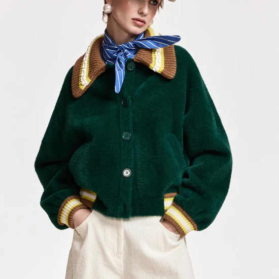A model wearing a dark green cropped button-up cardigan with a brown and yellow striped knit collar and cuffs, paired with cream trousers, a blue striped neck scarf, and a beige baseball cap.