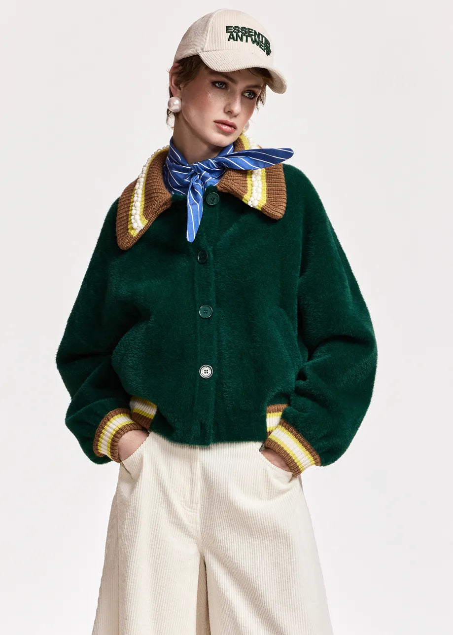 A model wearing a dark green cropped button-up cardigan with a brown and yellow striped knit collar and cuffs, paired with cream trousers, a blue striped neck scarf, and a beige baseball cap.