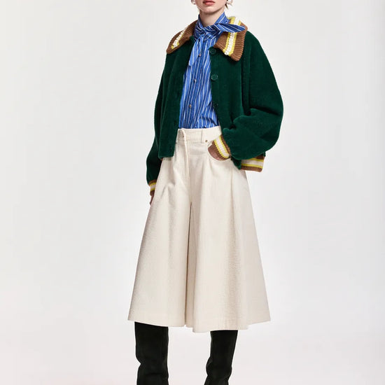 A model stands against a light gray studio background wearing a dark green cropped jacket with gold trim, a blue striped shirt, a cream wide-leg skirt, black knee-high boots, and a cream baseball cap.