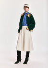 A model stands against a light gray studio background wearing a dark green cropped jacket with gold trim, a blue striped shirt, a cream wide-leg skirt, black knee-high boots, and a cream baseball cap.