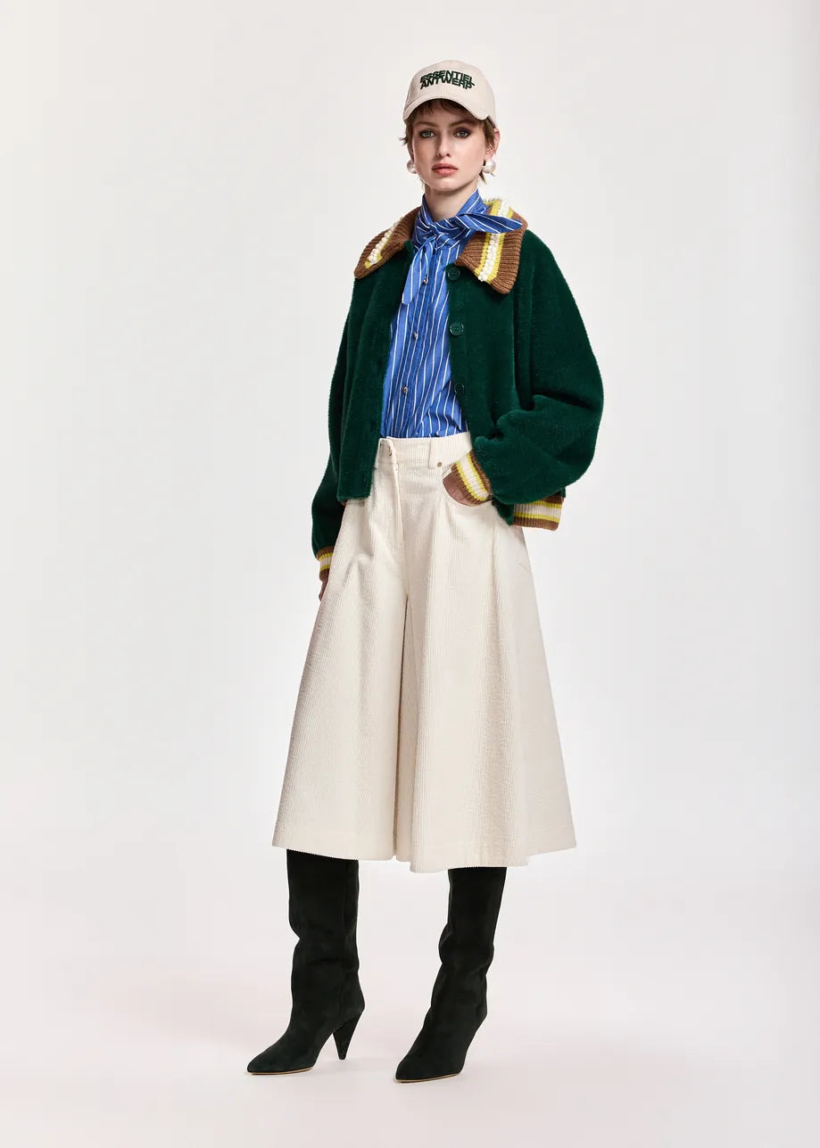 A model stands against a light gray studio background wearing a dark green cropped jacket with gold trim, a blue striped shirt, a cream wide-leg skirt, black knee-high boots, and a cream baseball cap.