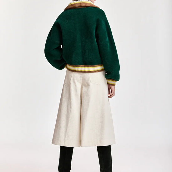 A model is shown from the back wearing a dark green cropped knit cardigan with gold and cream ribbed trim, a cream wide-leg skirt, black boots, and a cream baseball cap, set against a plain light gray studio background.