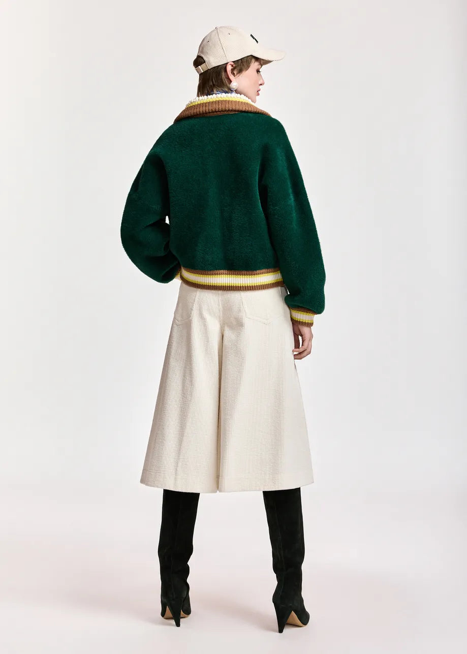A model is shown from the back wearing a dark green cropped knit cardigan with gold and cream ribbed trim, a cream wide-leg skirt, black boots, and a cream baseball cap, set against a plain light gray studio background.