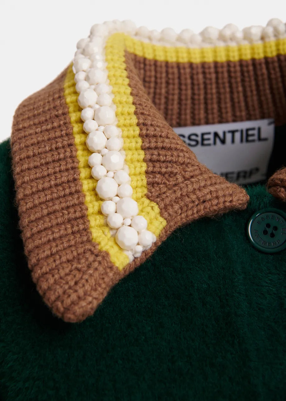 Close-up of a knit cardigan collar with brown and yellow stripes, white pom-pom trim, and a dark green wool body with a visible button.