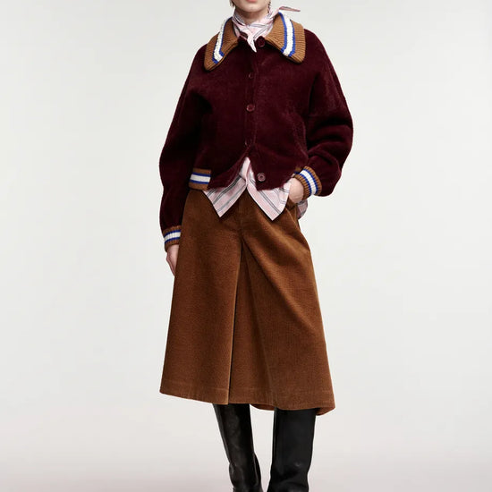 A model stands against a light gray studio background wearing a dark burgundy corduroy jacket with brown ribbed trim and striped cuffs, a brown midi skirt, black knee-high boots, a patterned scarf, and a tan cap.