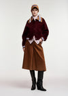 A model stands against a light gray studio background wearing a dark burgundy corduroy jacket with brown ribbed trim and striped cuffs, a brown midi skirt, black knee-high boots, a patterned scarf, and a tan cap.