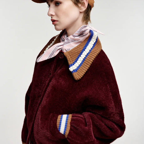 A model wearing a maroon plush fleece jacket with a striped knit collar and cuffs, a pink striped shirt, a pink neck scarf, and a brown baseball cap, photographed against a light gray studio background.
