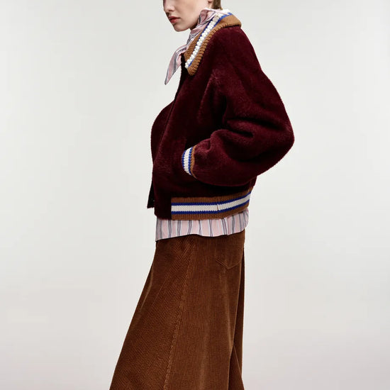 A model is shown from the side wearing a maroon fuzzy cropped jacket with striped ribbed trim, a brown corduroy skirt, a striped shirt, a patterned neck scarf, and a brown baseball cap, set against a plain light gray studio background.