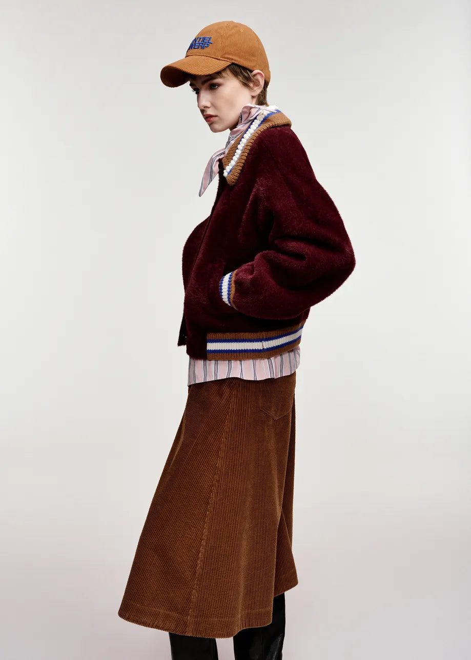 A model is shown from the side wearing a maroon fuzzy cropped jacket with striped ribbed trim, a brown corduroy skirt, a striped shirt, a patterned neck scarf, and a brown baseball cap, set against a plain light gray studio background.