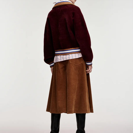 A model shown from the back wearing a dark burgundy fuzzy cropped cardigan over a striped shirt, a brown corduroy skirt, and black knee-high boots, against a plain light gray studio background.