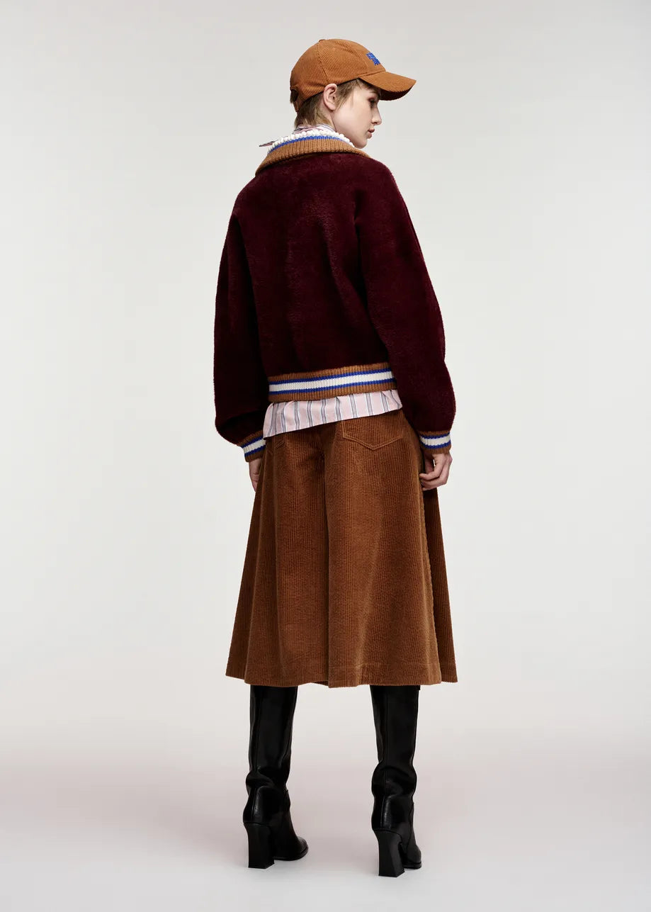 A model shown from the back wearing a dark burgundy fuzzy cropped cardigan over a striped shirt, a brown corduroy skirt, and black knee-high boots, against a plain light gray studio background.