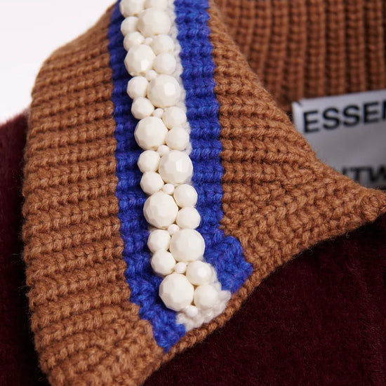 Close-up of a chunky knit sweater collar in brown and blue with a vertical row of white faux-pearl beads, resting on a burgundy fabric surface.