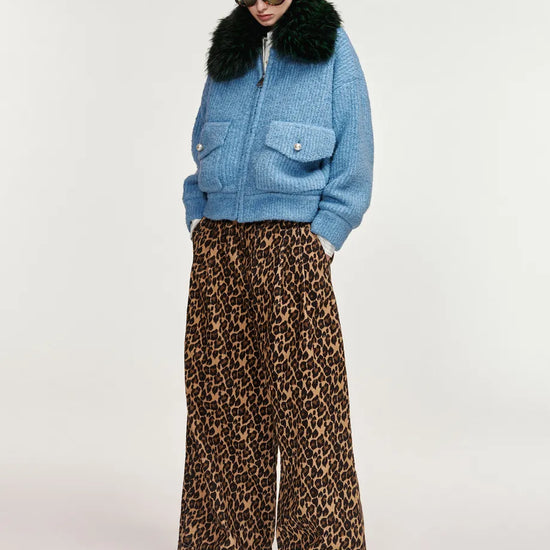 A model wearing a light blue textured jacket with a black fur collar, paired with wide-leg leopard print trousers, against a plain light gray studio background.