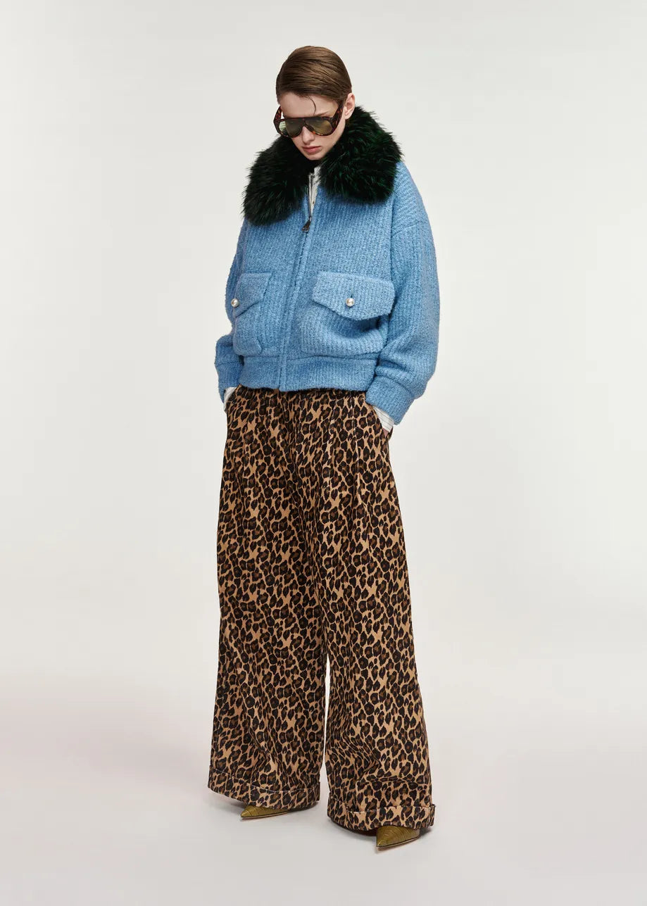 A model wearing a light blue textured jacket with a black fur collar, paired with wide-leg leopard print trousers, against a plain light gray studio background.