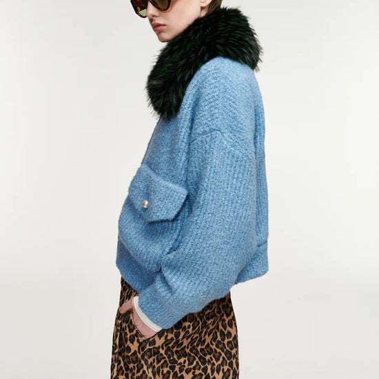 A model shown in profile wearing a light blue textured knit sweater with a dark green faux-fur collar, paired with a leopard-print skirt and tortoiseshell sunglasses, against a plain light gray studio background.