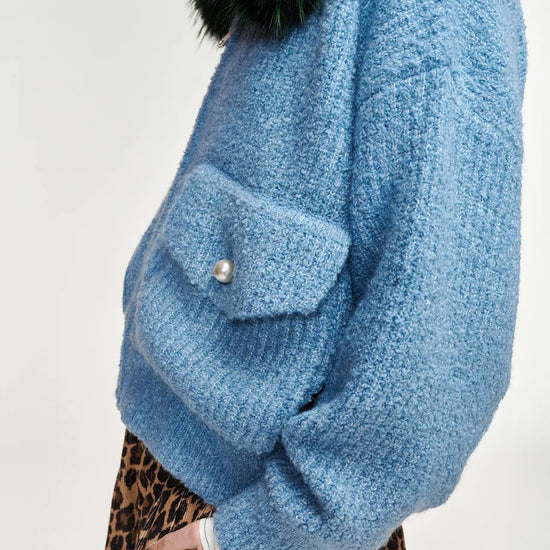 A close-up side view of a person wearing a chunky light blue knit sweater with a dark green faux-fur collar and a leopard-print skirt, against a plain off-white background.