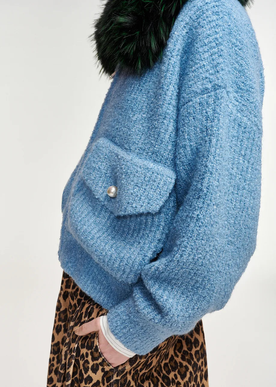 A close-up side view of a person wearing a chunky light blue knit sweater with a dark green faux-fur collar and a leopard-print skirt, against a plain off-white background.