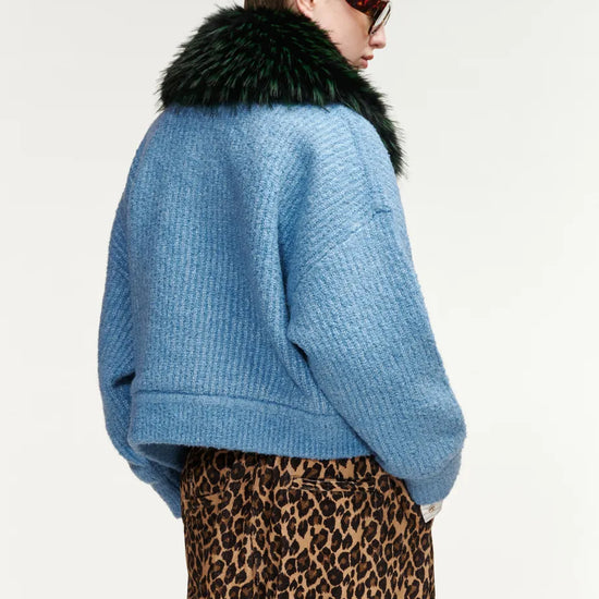 A model shown from the back wearing a light blue textured knit cropped jacket with a dark green faux-fur collar, paired with a leopard-print skirt, against a plain white studio background.