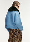 A model shown from the back wearing a light blue textured knit cropped jacket with a dark green faux-fur collar, paired with a leopard-print skirt, against a plain white studio background.
