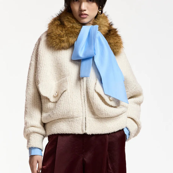 A model wearing a cream-colored textured jacket with a brown fur collar, a light blue scarf tied in a bow, and dark burgundy wide-leg trousers, set against a plain light gray background.