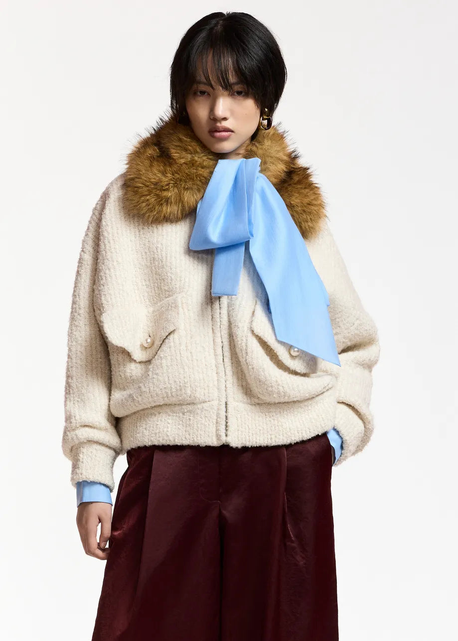 A model wearing a cream-colored textured jacket with a brown fur collar, a light blue scarf tied in a bow, and dark burgundy wide-leg trousers, set against a plain light gray background.