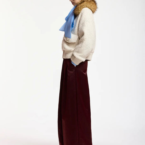 A model is shown from a side profile wearing a cream-colored textured knit sweater with a brown fur-trimmed collar, a light blue scarf, and wide-leg burgundy trousers, set against a plain white studio background.