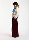 A model is shown from a side profile wearing a cream-colored textured knit sweater with a brown fur-trimmed collar, a light blue scarf, and wide-leg burgundy trousers, set against a plain white studio background.