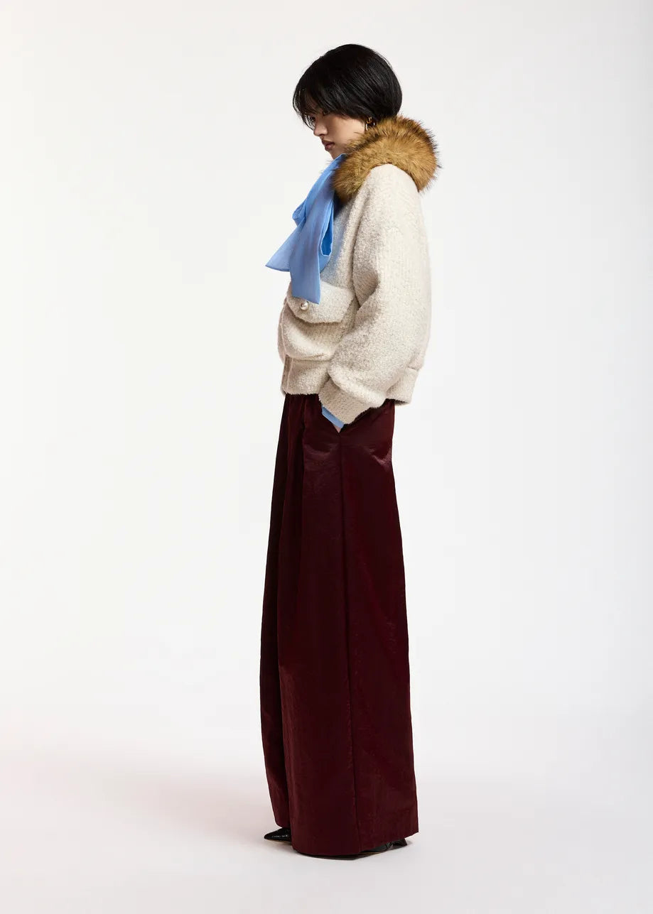 A model is shown from a side profile wearing a cream-colored textured knit sweater with a brown fur-trimmed collar, a light blue scarf, and wide-leg burgundy trousers, set against a plain white studio background.