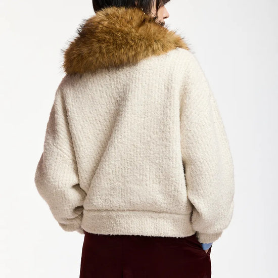 A person shown from the back wearing a cream sherpa fleece jacket with a large brown faux-fur collar, paired with dark burgundy pants, against a plain light gray studio background.