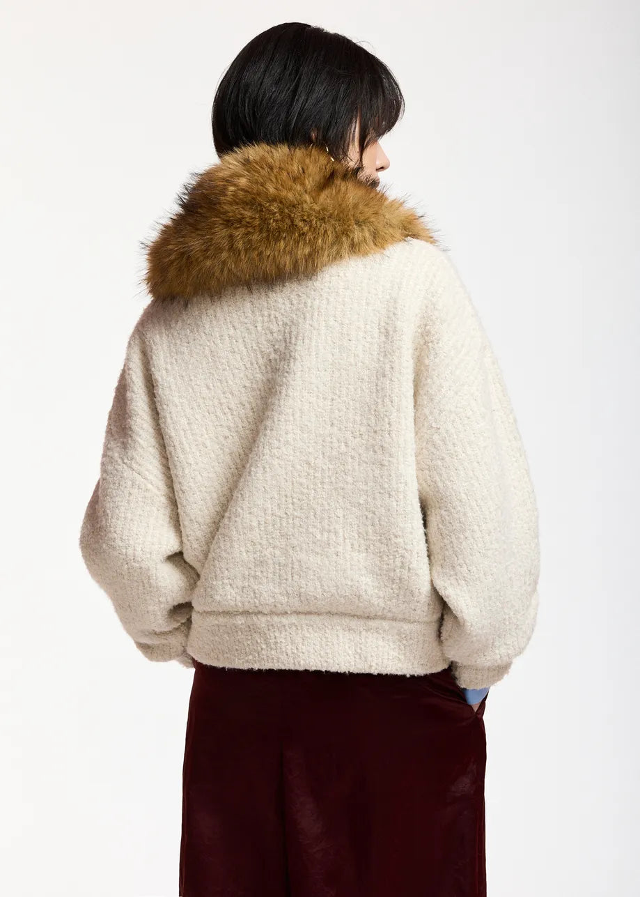 A person shown from the back wearing a cream sherpa fleece jacket with a large brown faux-fur collar, paired with dark burgundy pants, against a plain light gray studio background.